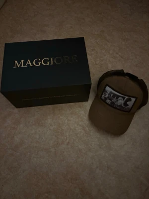 Brun Maggiore keps med fototryck - Säljer en brun keps från Maggiore med ett svartvitt fototryck framtill. Kepsen har klassisk böjd skärm och justerbar rem bak. Perfekt för dig som vill ha en unik accessoar med streetwear-vibe.