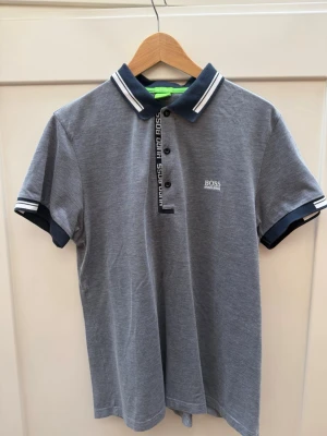 Grå pikétröja från Hugo Boss - Snygg grå pikétröja från Hugo Boss med vita och marinblå detaljer på krage och ärmslut. Tröjan har knappslå med logotyp, korta ärmar och är tillverkad i mjuk bomull. Slim fit-modell som ger en modern siluett. Knappt använd 