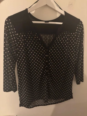 Svart prickig blus med trekvartsärm - Snygg svart blus från H&M med vita prickar och trekvartsärm. Blusen har knappar framtill och en lätt v-ringning. Materialet är tunt och luftigt, perfekt för lager på lager. Klassisk och tidlös design som passar till mycket.