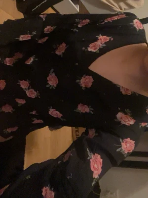 Svart blommig klänning med trumpetärm - Supersnygg svart klänning med rosa blommönster och trendiga trumpetärmar. Klänningen har en cool cut-out detalj vid axeln och är gjord i ett lätt, flowy tyg som ger en skön känsla. Perfekt för dig som gillar romantiska och feminina plagg med en twist.