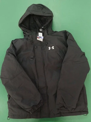 Svart vindjacka från Under Armour - Svart vindjacka från Under Armour med huva och dragsko. Jackan har dragkedja framtill, två bröstfickor och elastiska muddar vid ärmsluten. Materialet är tåligt och vattenavvisande, perfekt för blåsiga dagar. Under Armour-logga i vitt på bröstet. Tagsen är borta dock