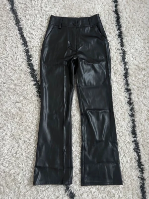 Svarta fuskskinnbyxor med raka ben - Snygga svarta byxor från Madlady i storlek XS i fuskskinn med hög midja. Byxorna har bälteshällor, knapp och dragkedja framtill samt fickor på sidorna. Perfekta för en trendig look och passar till många olika stilar.