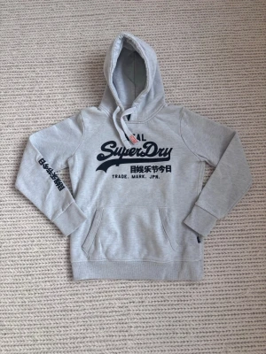 Grå Superdry hoodie med tryck - Snygg grå hoodie från Superdry med svart logga och japanska tecken på bröstet och ärmen. Nyskick. Storlek S