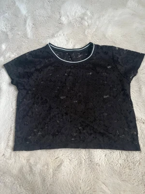 Svart spetsig croppad topp - Säljer en svart croppad topp i spets med korta ärmar och rund halsringning med vit kant. Perfekt för att styla lager på lager eller bära som den är för en edgy look.