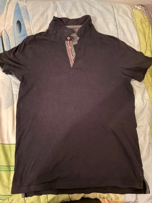 Svart kortärmad polotopp med knappar - Svart polotopp med korta ärmar och unik knappdetalj vid halsen. Toppen har en hög krage som kan bäras uppvikt eller nedvikt och är tillverkad i mjukt bomullsmaterial. Passar perfekt till jeans eller kjol för en clean och stilren look.