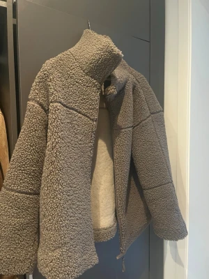 Grå teddyjacka med dragkedja - Mysig grå teddyjacka i oversized modell med hög krage och dragkedja framtill. Jackan har breda ärmar och ett fluffigt, fårskinnsinspirerat material som ger en cool och avslappnad look. 