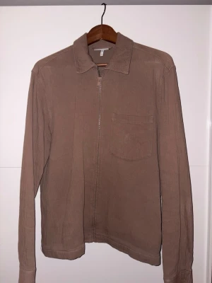 Beige overshirt med dragkedja - Stilren beige overshirt med dragkedja framtill och en bröstficka. Klassisk krage och långa ärmar med knapp vid ärmslut. Tillverkad i ett mjukt tyg som ger en avslappnad look. Perfekt att bära över en t-shirt för en enkel och snygg stil.