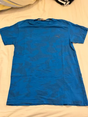 Blå camo t-shirt från Puma - Snygg blå t-shirt från Puma med diskret camouflagemönster och klassisk rund hals. T-shirten har korta ärmar och är tillverkad i ett mjukt bomullsmaterial som känns skönt mot huden. Men storleken är liten Storlek passar till M 