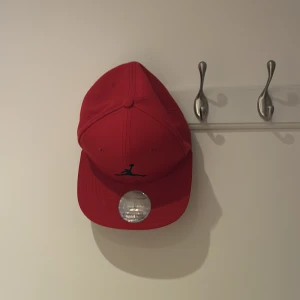 Röd snapback keps från Jordan - Snygg röd snapback keps från Jordan med klassisk logga broderad framtill. Kepsen har platt skärm och justerbar baksida för perfekt passform. Insidan har svarta detaljer. Perfekt för en sportig och trendig look.