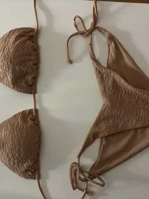Beige bikini med struktur och knyt - Trendig beige bikini med strukturerat tyg och klassisk trekantsmodell. Både överdel och underdel har knytband för justerbar passform. Perfekt för stranden eller poolen under sommaren. Använd fåtal gånger men såklart tvättat så den är som ny. 