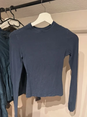Mörkblå långärmad topp - En enkel och stilren mörkblå långärmad topp med rund hals. Modellen är croppad och har en tight passform som sitter snyggt på kroppen. Perfekt att matcha med jeans eller kjol för en clean look.