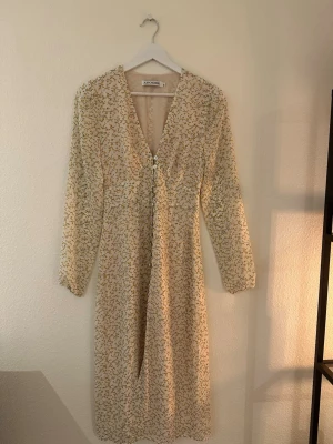 Blommig långklänning från Adoore - Söt långklänning från Adoore i ljusbeige med små blommor i gult och grönt. Klänningen har v-ringning, knappar framtill och långa ärmar. Perfekt för sommaren! 