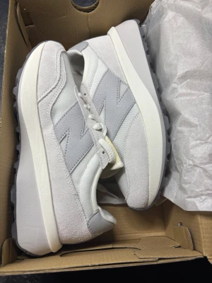 New Balance 370 - Snygga sneakers från New Balance i vitt, beige och ljusgrått med klassisk N-logga på sidan. Ovandelen är i en mix av mocka och mesh, vilket ger både stil och andningsförmåga. Tjock sula för extra komfort och cool retrovibe. Perfekta för dig som gillar streetstyle.