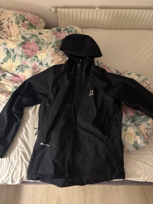 Svart vindjacka från Haglöfs Gore-Tex - Snygg svart vindjacka från Haglöfs med huva och dragkedja framtill. Jackan är tillverkad i slitstarkt Gore-Tex-material och har flera praktiska fickor med dragkedja. Perfekt för blåsiga dagar och outdoor-äventyr.