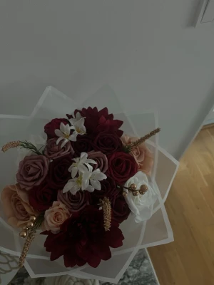 Bukett med konstgjorda blommor - Säljer en vacker bukett med konstgjorda blommor i vinrött, rosa, vitt och guld. Buketten har rosor, liljor och dekorativa gulddetaljer, inslagen i transparent papper. Perfekt som dekoration eller present.