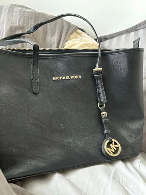 Svart handväska från Michael Kors - Snygg svart handväska från Michael Kors i strukturerat läder med guldfärgade detaljer och MK-hänge. Väskan har dragkedja upptill, flera innerfack och ljusbeige foder. Perfekt för dig som vill ha en stilren och rymlig väska med exklusiv känsla.banden är lite lite trasiga men inget som påverkad väskan!