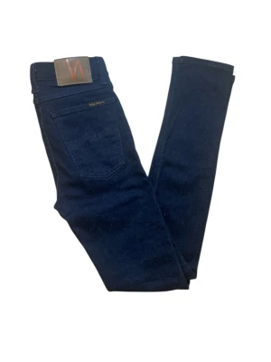 Mörkblå  jeans från Nudie - Snygga mörkblå skinny jeans från Nudie Jeans med klassisk femficksdesign och läderpatch bak i midjan. Jeansen har smal passform och är tillverkade i stretchig denim för extra komfort. Perfekta för dig som gillar en stilren och enkel look. Superbra skick 