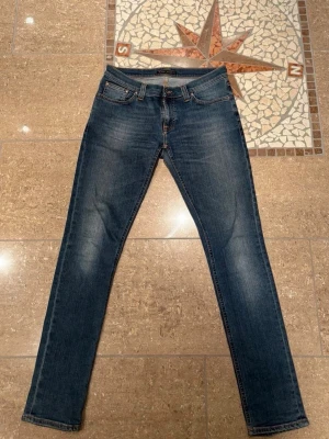 Blå jeans från Nudie Jeans - Säljer ett par klassiska blå jeans från Nudie Jeans med smal passform och snygga slitningar. Dom är sparsamt använda, inga tecken på användning och inga hål. Storlek 30/32.
