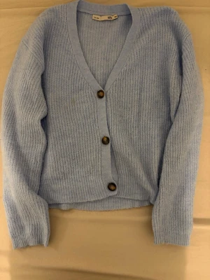 Ljusblå ribbad kofta med knappar - Mysig ljusblå kofta från H&M med ribbad struktur och djup V-ringning. Koftan har tre stora bruna knappar framtill och långa ärmar. Perfekt att slänga på sig för en chill look. Har en fläck.