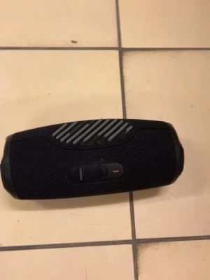 Svart JBL bärbar högtalare - Snygg svart bärbar högtalare från JBL med robust tygmaterial och gummidetaljer. Cylindrisk form med tydliga knappar och diskret JBL-logga på sidan. Perfekt för att ta med sig överallt och spela musik trådlöst via Bluetooth.