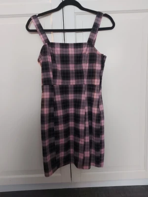 Rutig rosa och svart klänning - Snygg kort klänning med rutigt mönster i rosa och svart. Klänningen har breda axelband och dragkedja i sidan för enkel påklädning. Perfekt för dig som gillar en trendig och ungdomlig stil.