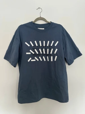 Blå Maison Margiela t-shirt - Original Maison Margiela T-shirt i blå/grå nyans. Ikonisk Margiela-design med numrerad print framtill och de klassiska fyra vita stygnen baktill vid nacken. 	•	Storlek: 48 (passar M/L beroende på fit) 	•	Tillverkad i Italien 	•	Skick: Mycket bra, inga hål eller fläckar 	•	100 % bomull 	•	Unisex  Tidlös designer-tee som funkar lika bra casual som stylad. Skickas snabbt 📦 