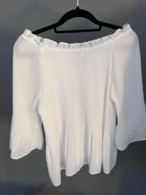 Vit offshoulder blus med volang - Säljer en superfin vit offshoulder blus med volangkant upptill och vida trekvartsärmar. Blusen är lätt plisserad och har en luftig, flowy passform som ger en romantisk vibe. Perfekt till jeans eller kjol för en trendig look.
