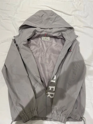 Grå vindjacka från Moncler - Snygg grå vindjacka från Moncler med huva och dragkedja framtill. Jackan har elastiska muddar vid ärmslut och nederkant, samt Moncler-logga på ärmen. Tillverkad i lätt och tåligt material, perfekt för blåsiga dagar. Det är bara att höra av sig vid några frågor, kan fixa mer bilder om de också behövs