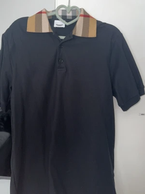 Svart piké med mönstrad krage Burberry - Snygg svart pikétröja från Burberry med klassisk rutmönstrad krage i beige, rött och svart. Tröjan har korta ärmar och knäppning med tre knappar framtill. Perfekt för en stilren och trendig look.