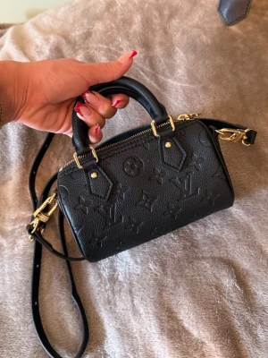 Louis Vuitton svart mini handväska - Nano Speedy - Säljer en svart mini handväska från Louis Vuitton i präglat läder med klassiskt monogrammönster. Väskan har både handtag och juster sopbar axelrem i svart läder samt guldfärgade metalldetaljer. Perfekt för dig som gillar lyxiga accessoarer med ikonisk design.