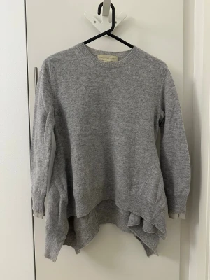 Grå stickad tröja Stella McCartney - Snygg grå stickad tröja från Stella McCartney med rund halsringning och asymmetrisk nederkant. Tröjan har långa ärmar och är tillverkad i mjukt material, perfekt för lager på lager och en avslappnad men trendig look.