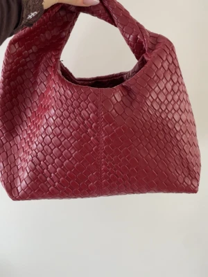 Röd flätad handväska  - Snygg röd handväska med flätad design och mjuk, rundad form. Väskan är tillverkad i skinn liknande material och har ett kort handtag som gör den enkel att bära i handen. Perfekt accessoar för att lyfta din outfit