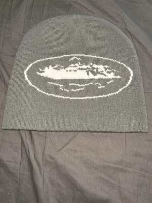 Cortez mössa - Säljer en stilren grå mössa med vit broderad logga framtill. Mössan är ribbstickad och har en klassisk, rundad form som sitter skönt på huvudet. Perfekt accessoar för kyliga dagar och enkel att matcha med olika outfits.