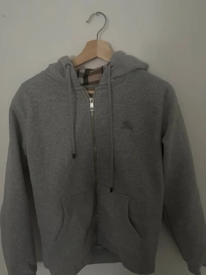 Grå zip hoodie från Burberry - Snygg grå hoodie med dragkedja från Burberry. Klassisk huva med dragsko och fickor framtill. Insidan av huvan har det ikoniska Burberry-rutiga mönstret. Tillverkad i mjukt bomullsmaterial, perfekt för en avslappnad stil.