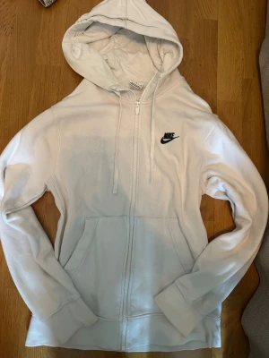 Vit zip hoodie från Nike - Säljer en clean vit hoodie från Nike med dragkedja framtill och klassisk huva med dragsko. Nike-logga i svart broderad på bröstet. Mjuk bomullsblandning och två stora fickor framtill. Passar för både killar och tjejer