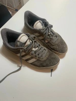 Grå Adidas sneakers i mocka - Snygga grå sneakers från Adidas med klassiska vita ränder på sidan. Skorna är låga och har ovandel i mocka samt vita detaljer på plösen.  I bra skick men smutsiga.