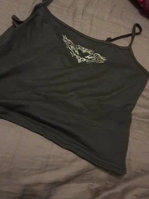 Svart croppad linnetopp med strassdetalj - Snygg svart croppad linnetopp med tunna axelband och en glittrig strassdetalj framtill. Perfekt att styla med jeans eller kjol för en trendig look. Enkel och stilren design som passar till många outfits.