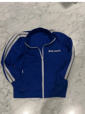 Blått tracksuit set från Palm Angels - Säljer ett blått tracksuit set från Palm Angels med vit dragkedja och vita ränder längs ärmar och ben. Setet består av en zip-jacka med hög krage och matchande byxor med resår i midjan. Båda delarna har Palm Angels-logga tryckt i vitt. Passar både M och S 