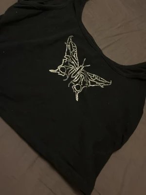 Svart croppad topp med fjärilsdetalj - Snygg svart croppad topp med en stor fjäril framtill i glittriga stenar. Toppen har bred urringning och är ärmlös, perfekt för att sticka ut lite extra. Passar till många olika stilar och är lätt att matcha.