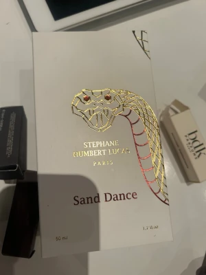 Sand Dance parfym Stephane Humbert Lucas - Lyxig parfym Sand Dance från Stephane Humbert Lucas Paris. Förpackningen är vit med en elegant guld- och rödfärgad orm i relief och text i guld. Flaskan rymmer 50 ml och designen känns exklusiv och konstnärlig. Nästan helt full