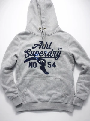 Grå hoodie från Superdry - Snygg grå hoodie från Superdry med stor blå logga och tryck framtill. Klassisk känguruficka och dragsko i huvan. Mjuk insida och ribbade muddar. Perfekt för en avslappnad streetstyle-look.