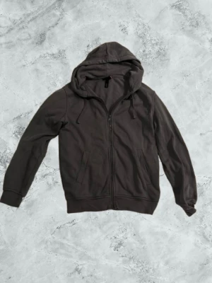 Mörkgrå zip-hoodie med huva - En stilren mörkgrå hoodie med dragkedja och huva. Modellen har två fickor framtill och justerbara snören i huvan. Perfekt för en avslappnad och cool look. Passar till det mesta och är enkel att matcha med andra plagg.