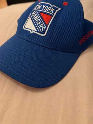 Blå New York Rangers keps Reebok - Snygg blå keps med New York Rangers-logga broderad framtill. Kepsen är från Reebok och har röda detaljer samt böjd skärm. Perfekt för dig som gillar hockey och vill visa vilket lag du hejar på.