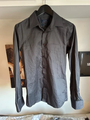 Mörkgrå skjorta från J.F.K - Stilren mörkgrå skjorta från J.F.K med klassisk krage och bröstficka. Skjortan har långa ärmar, knappar framtill och är tillverkad i ett slätt material som ger en snygg look. Perfekt för dig som gillar en enkel men elegant stil.