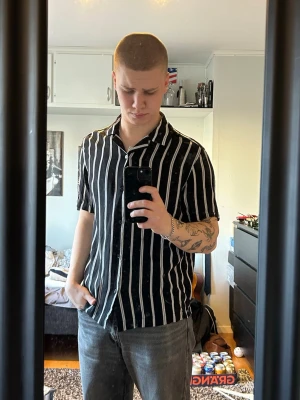 Svartvit randig skjorta med knappar - Säljer en svart skjorta med vita vertikala ränder och knappar framtill. Skjortan har korta ärmar och klassisk krage, perfekt för en avslappnad och cool stil. Materialet känns lätt och bekvämt, idealiskt för varmare dagar.