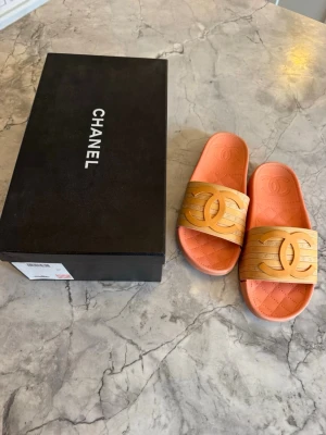 Chanel slides med logga - Chanel slides/badsandaler i storlek 37. Jag har vanligtvis storlek 38-38,5 cm. Har passat mig utmärkt i storlek Perfekta for sommar, semester och vardag. Saljer mina Chanel sandaler i en snygg korallfärgad ton med beige och guldfärgad CC-logga. De ar köpta i butik och levereras med originalkartong. Notera att det finns en defekt, sista bilden, på ena sandalen där den spruckit i gummit. Därav lågt pris Detaljer: • Klassisk quiltad sula med CC-detalj • Tydlig logga på både rem och undersida