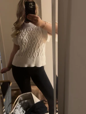 Vit kabelstickad topp med kort ärm - Snygg vit kabelstickad topp med korta ärmar och hög ribbad krage. Tröjan har ett strukturerat mönster framtill och breda ribbade muddar vid ärmslut och nederkant. Perfekt att styla med jeans eller kjol för en trendig look.