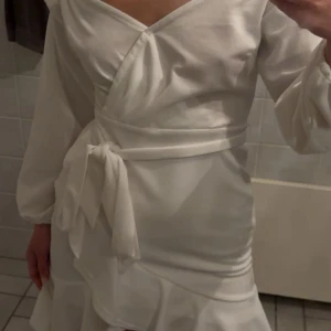 Student klänning/ vit klänning  - Elegant vit omlottklänning med offshoulder-design och volangdetaljer. Klänningen har långa ärmar och ett knytband i midjan för en smickrande passform. Aldrig använd, passar s också. 