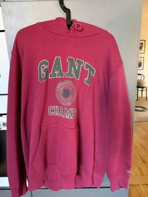 Röd hoodie från GANT med tryck - Snygg röd hoodie från GANT med grön och vit text på bröstet där det står 'GANT CHAMPS'. Klassisk känguruficka framtill, ribbade muddar och huva med dragsko. Perfekt för en avslappnad och sportig stil. Använd fåtal gånger