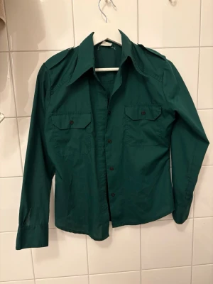 Mörkgrön skjorta med stora kragar - Snygg mörkgrön skjorta med stora retrokragar och två bröstfickor med knappar. Tillverkad i en mix av polyester och bomull, långärmad och med klassisk knappstängning framtill. Perfekt för dig som gillar vintagevibbar.